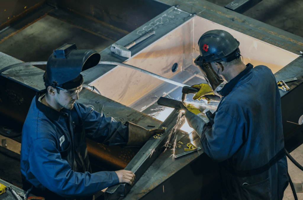 Two people wearing protective gear welding a piece of metal over a clear work table