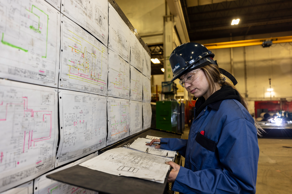 Engineer reviewing technical blueprints in an industrial facility, wearing safety gear and making notes on a clipboard.